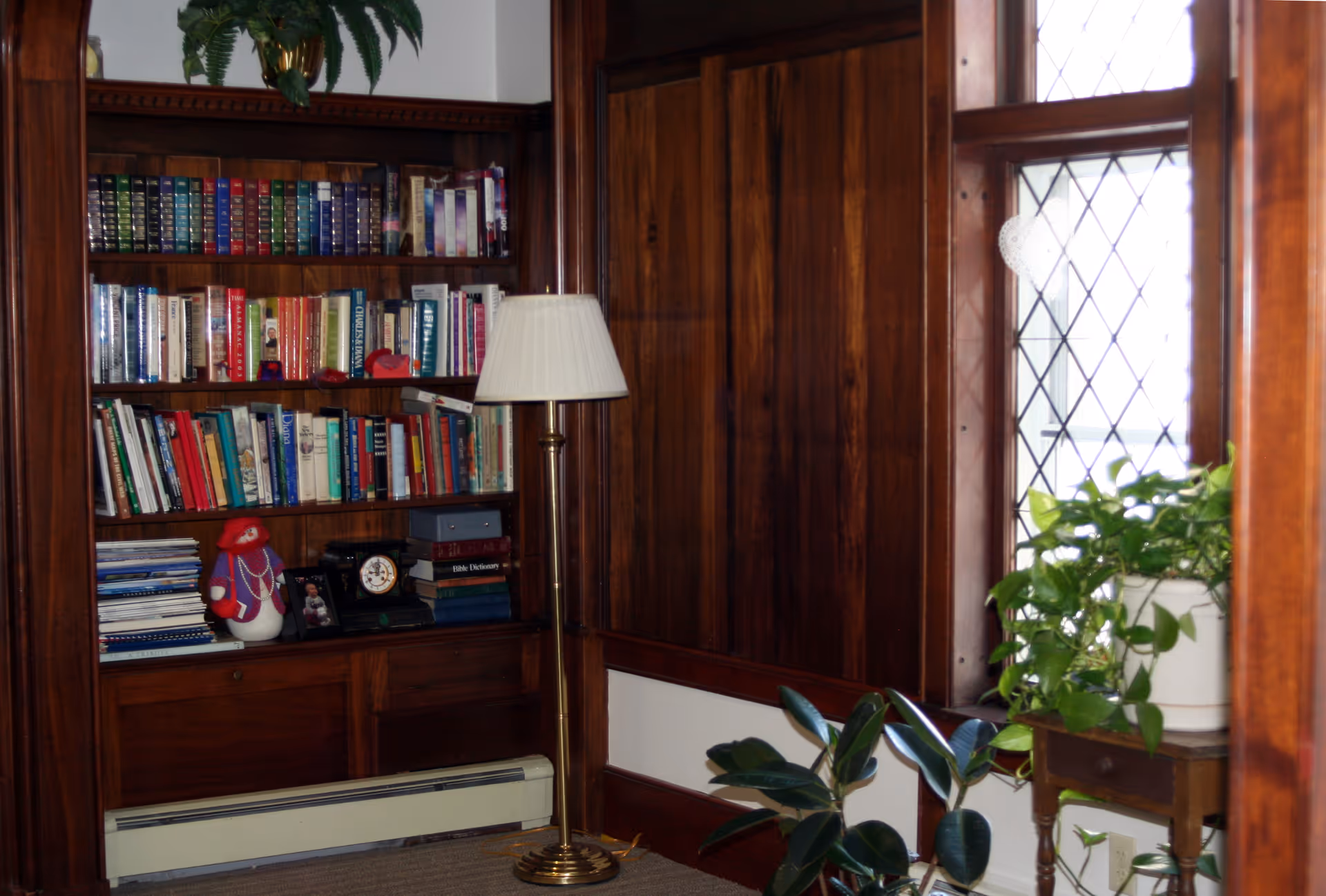 Wood-paneled reading nook with built-in bookshelves, a floor lamp, potted plants and a leaded glass window.