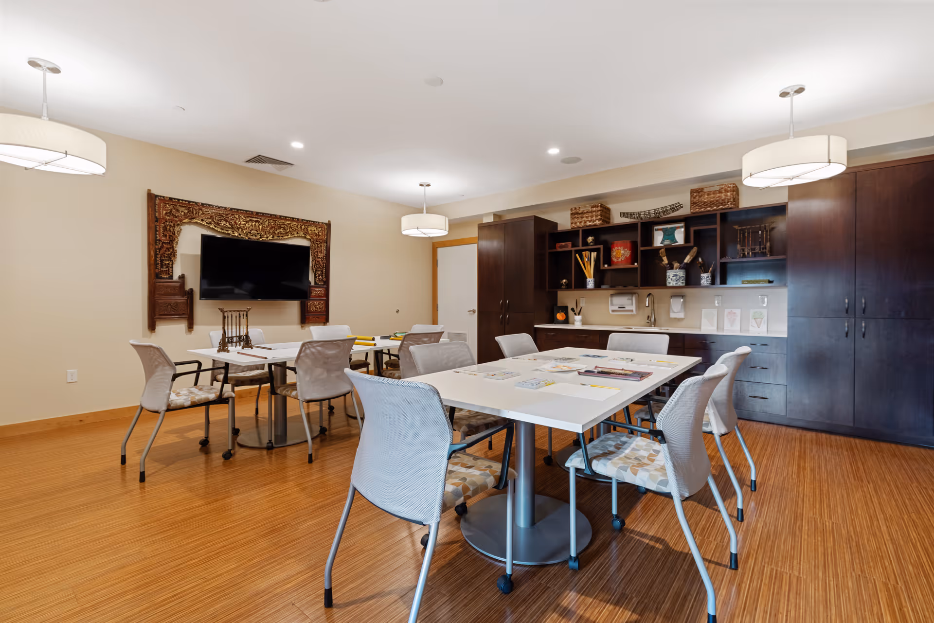 Bright communal activity/dining room with tables and chairs, a wall-mounted TV, and built-in cabinets.