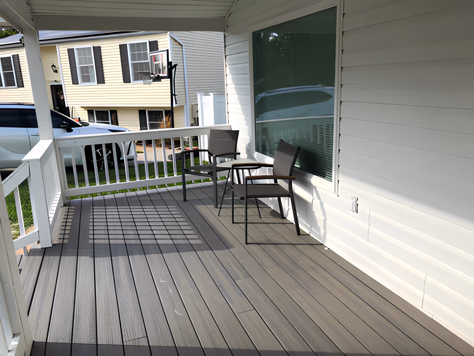 A covered outdoor porch area with two gray chairs and a small round table between them. The porch has gray wooden flooring and white railing. In the background, there is a view of a driveway with parked cars and a yellow house with black shutters.