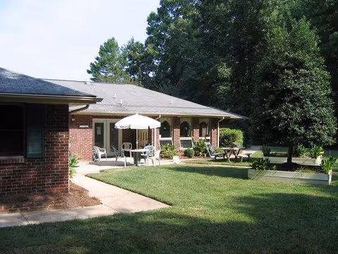 Brick senior living building with outdoor seating under umbrellas and a green lawn.