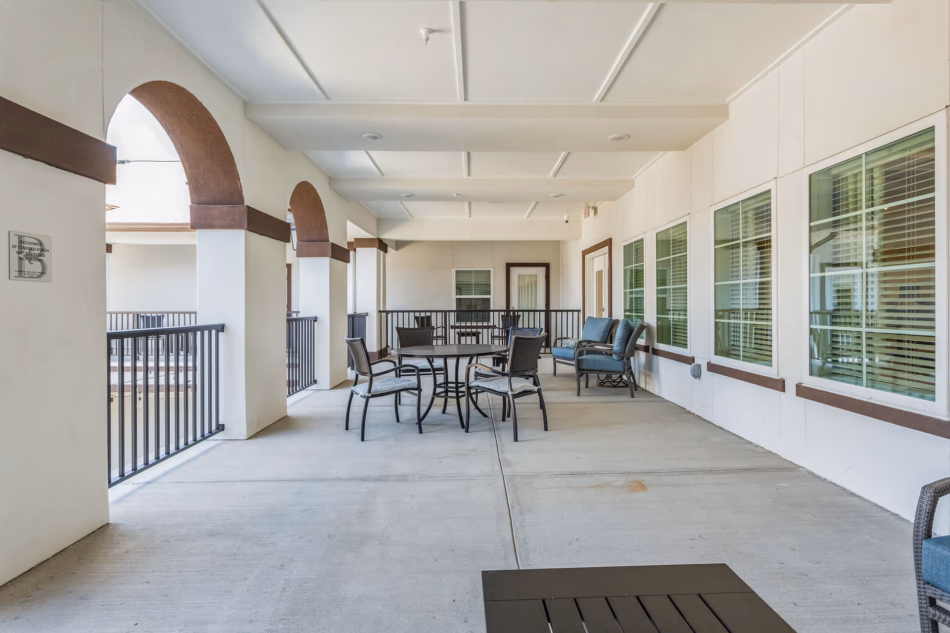 Covered outdoor seating area on a balcony with tables, chairs, arched openings, and windows along the building.