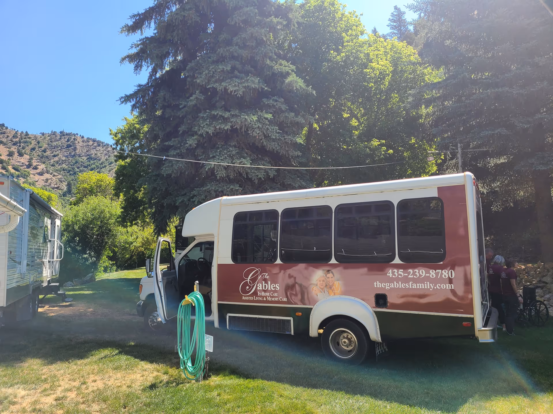 A small shuttle bus parked on grass near trees and a trailer. The bus has signage for The Gables Assisted Living of Brigham City, including a phone number and website. The bus door is open and a few people are visible near the back of the bus.