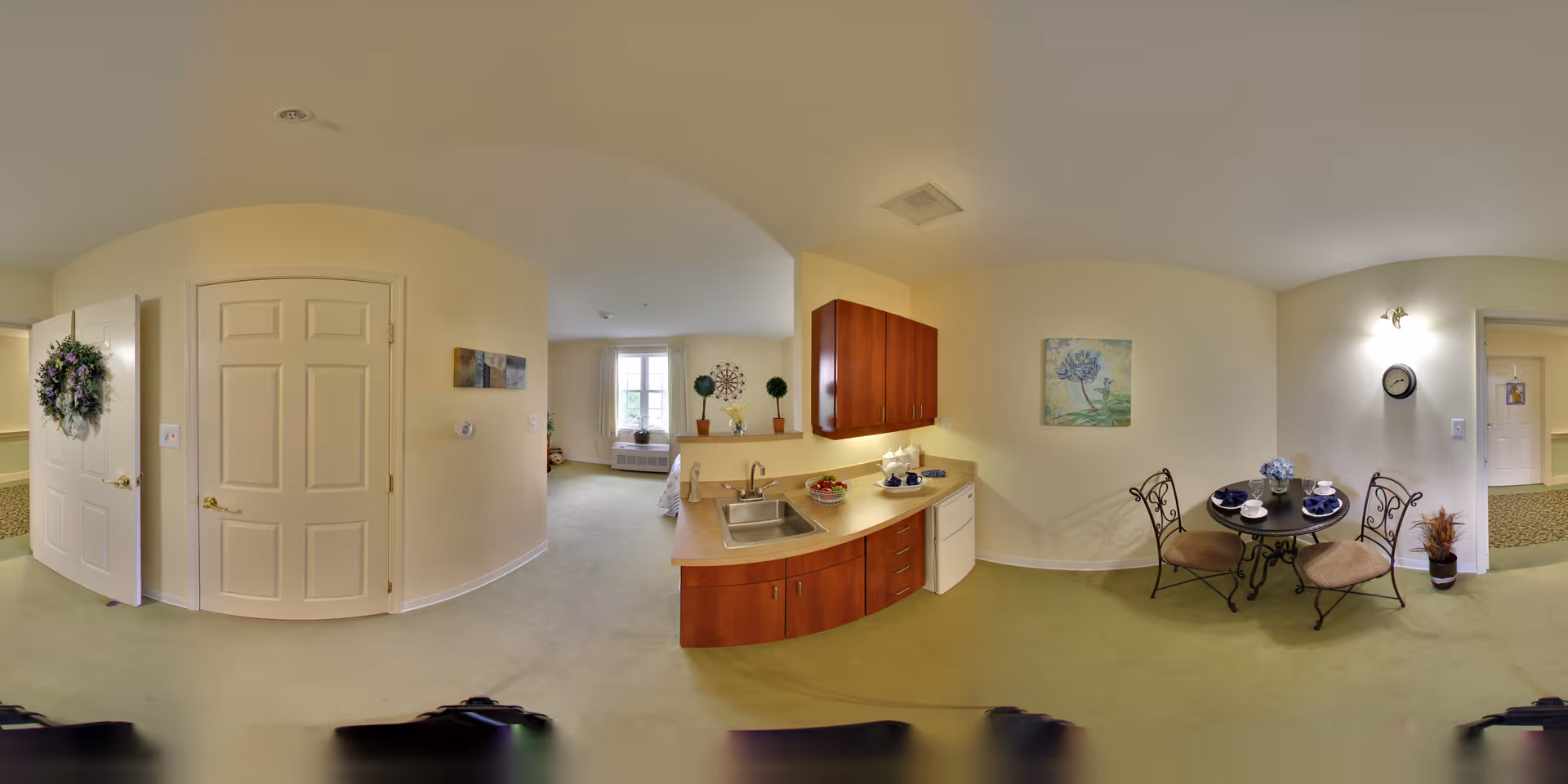 A cozy assisted living apartment interior featuring a small kitchen area with wooden cabinets, a sink, and a countertop. Adjacent to the kitchen is a round dining table set for two with chairs. The room has light yellow walls, a green carpet, and a window letting in natural light. There are decorative elements including a wall painting, a clock, and a potted plant.