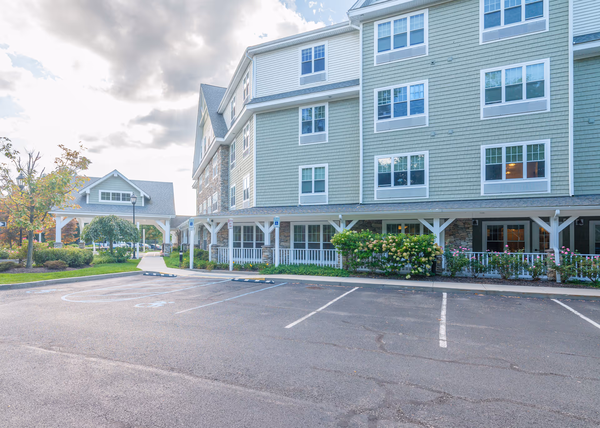 Exterior view of a senior living facility building with multiple windows, a covered walkway, landscaped bushes and trees, and an empty parking lot with marked spaces including handicapped spots.