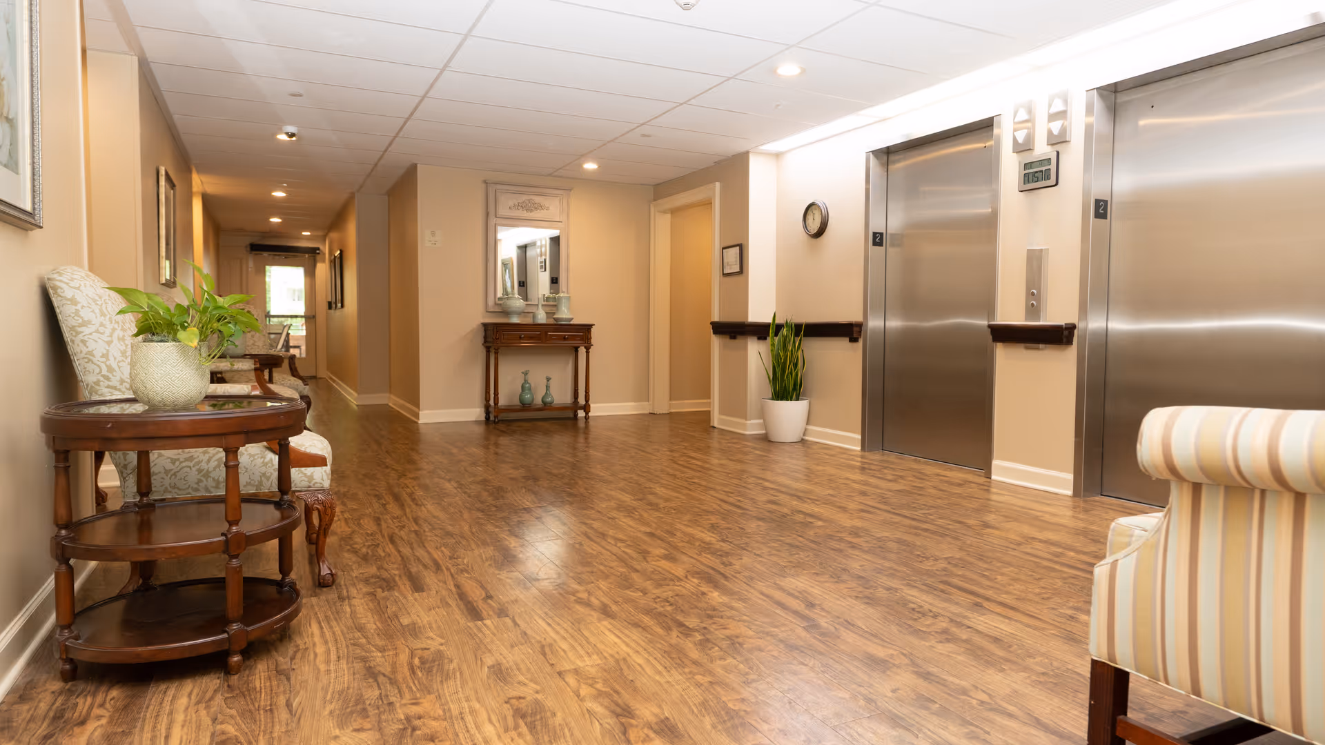 A well-lit hallway in a senior living facility with wooden flooring, two stainless steel elevators on the right, a small table with decorative vases and a mirror on the far wall, and comfortable chairs with a small round wooden table and a potted plant on the left side.