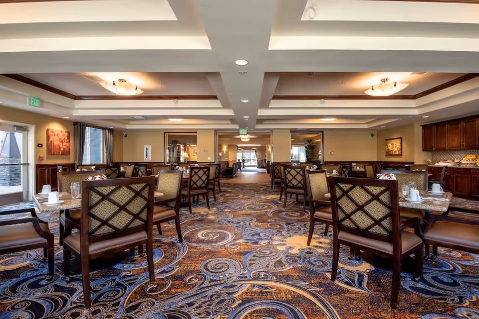 A spacious dining room in a senior living facility with multiple tables and chairs arranged neatly. The room features patterned carpet flooring, warm lighting from ceiling fixtures, and wooden cabinetry along one wall with a buffet setup. Large windows and glass doors allow natural light to enter the room.