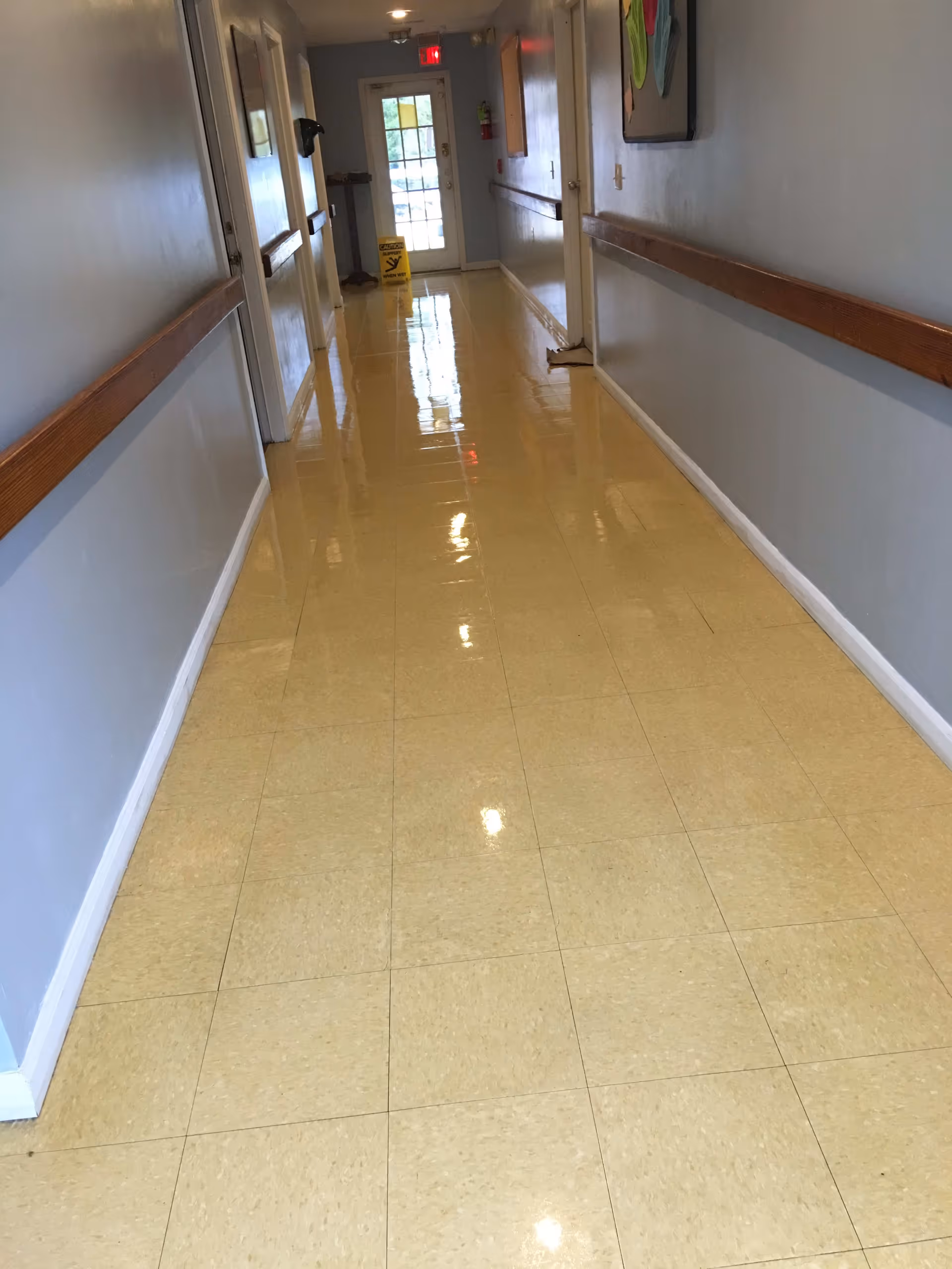 A clean, shiny hallway with beige tiled floor and light blue walls. Wooden handrails run along both sides of the hallway. At the end of the hallway, there is a glass door with a red exit sign above it and a yellow caution wet floor sign nearby. A fire extinguisher is mounted on the wall to the right of the door.