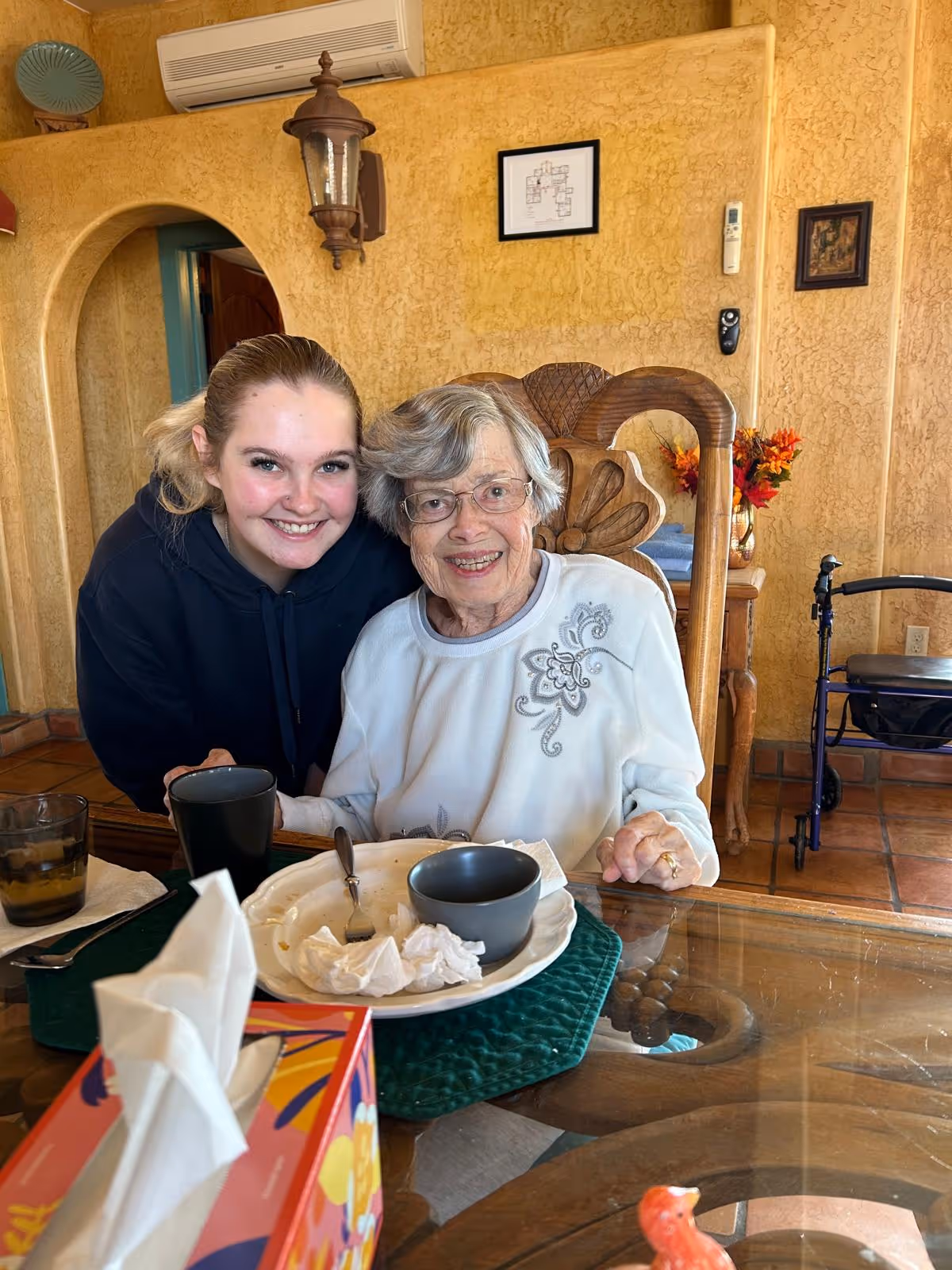 An elderly woman sitting at a dining table smiles with a younger woman leaning in beside her, plates and cups on the table and a walker nearby in a warm-toned dining room.
