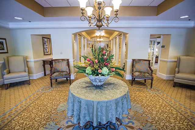A well-lit interior common area with a round table covered with a light blue patterned tablecloth and a large floral arrangement in the center. The room has patterned carpet flooring, beige walls with white trim, and several upholstered chairs arranged along the walls. A chandelier hangs from the ceiling, and a hallway with additional lighting and decor is visible in the background.