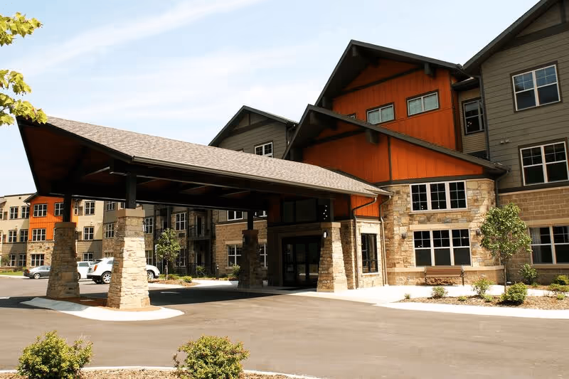Exterior view of a senior living facility building with a covered entrance supported by stone pillars, multiple windows, and a mix of orange and gray siding. There are small landscaped areas with bushes and a paved driveway in front.