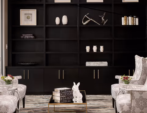 Stylish seating area with patterned armchairs around a glass coffee table in front of a large black built-in shelving unit with decorative objects.