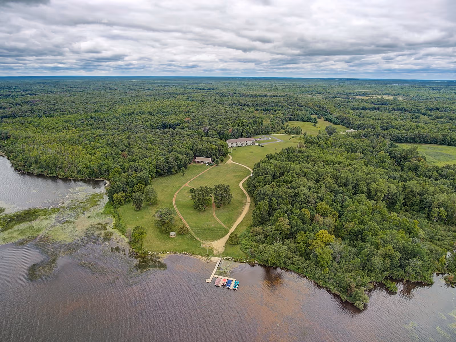 Aerial view of a senior living facility named MasonWoods surrounded by dense forest and a large body of water. The image shows a dock with several boats, a winding path leading to buildings, and open grassy areas under a cloudy sky.
