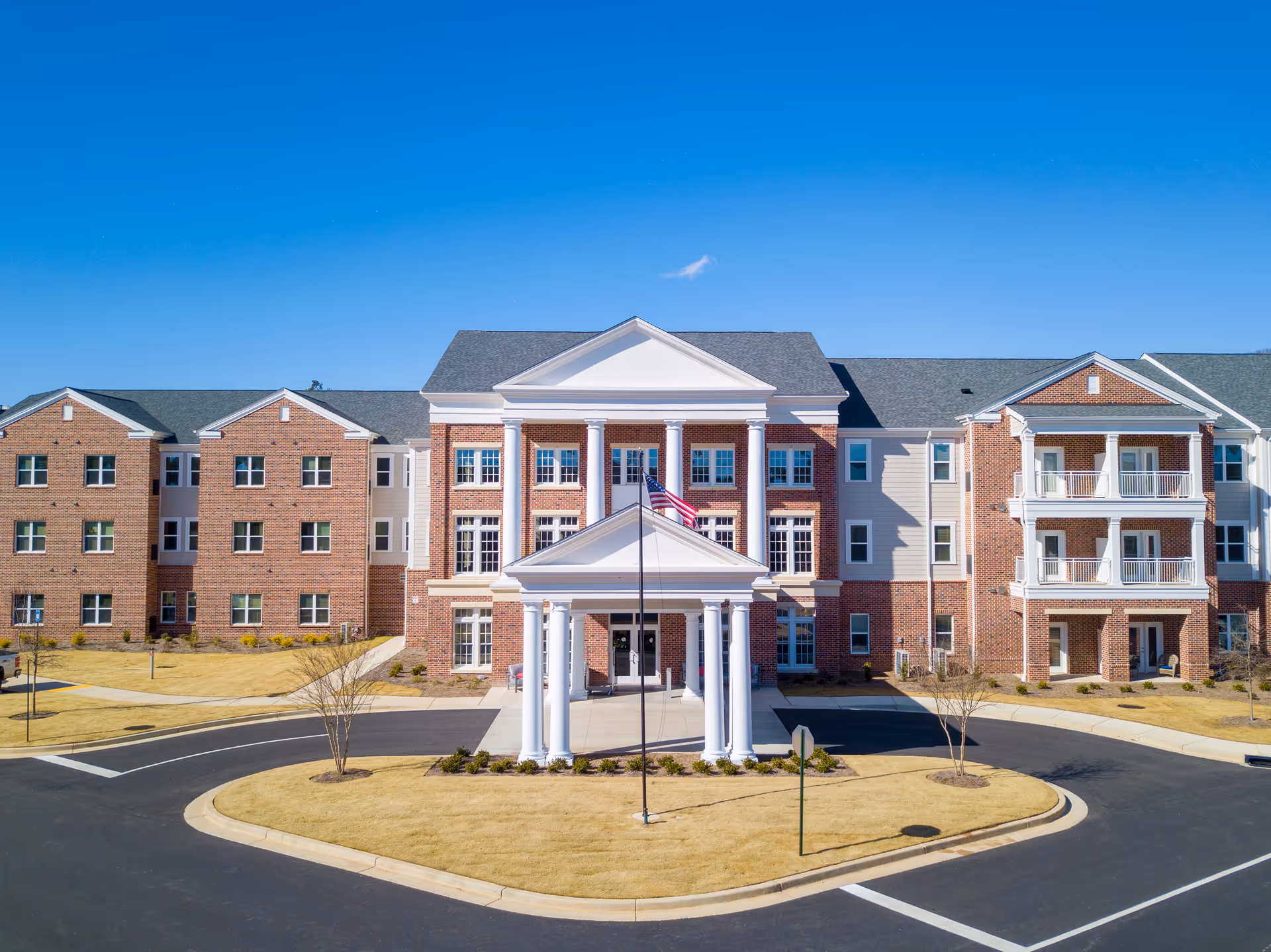 Front exterior view of Legacy Reserve at Old Town, a large brick and white-paneled senior living facility with a covered entrance supported by white columns, an American flag in front, and a circular driveway surrounded by landscaped grass and small trees under a clear blue sky.