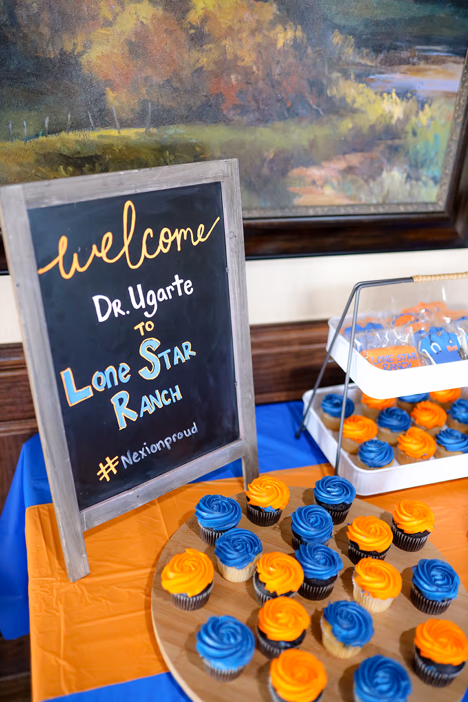 A welcome sign on a chalkboard reads 'Welcome Dr. Ugarte to Lone Star Ranch #Nexionproud' placed on a table with blue and orange tablecloths. The table also has a wooden tray with blue and orange frosted cupcakes and a two-tiered stand with more cupcakes and decorated cookies. A framed painting with autumn colors is visible in the background.
