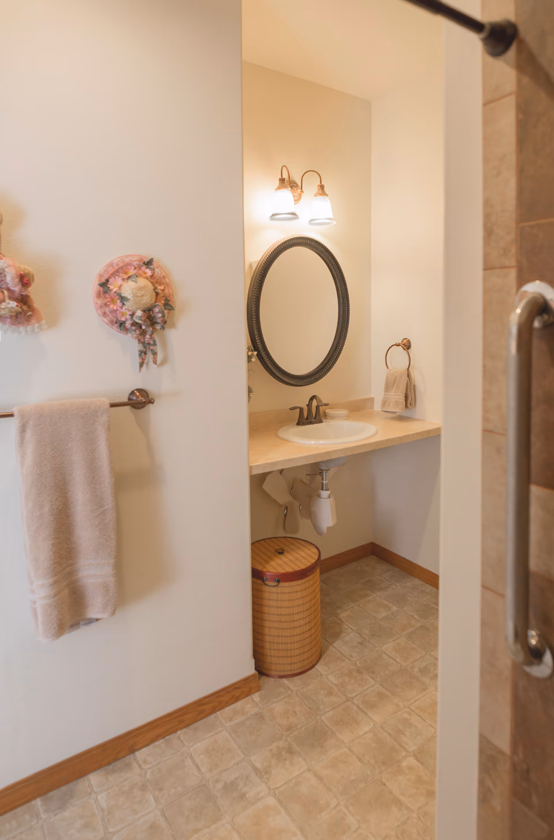 A bathroom with a beige tiled floor, a wall-mounted sink with a round mirror above it, two light fixtures, a towel hanging on a rack, decorative hats on the wall, and a wicker laundry basket under the sink.