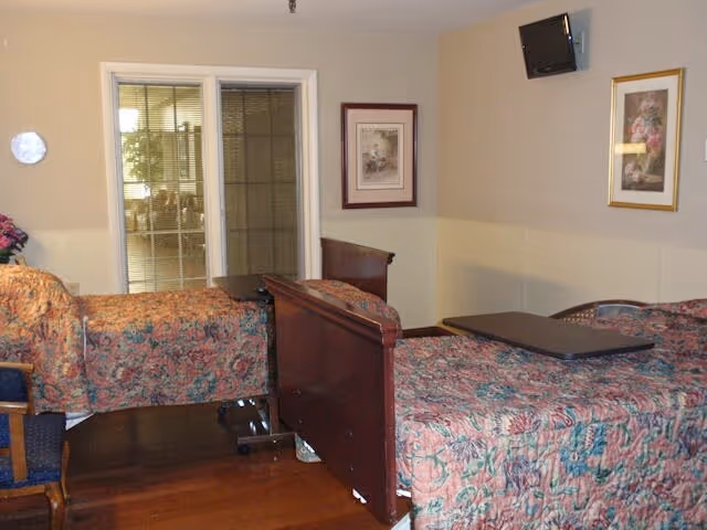 A room with two beds covered in floral patterned bedspreads, each with an overbed table. There is a wooden chair with blue upholstery, two framed pictures on the beige walls, a small wall-mounted TV, and a glass door leading to another room with natural light.