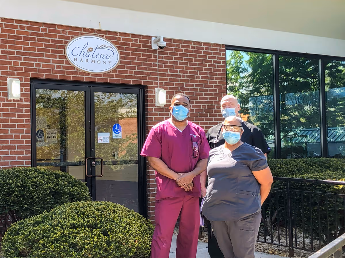 Three masked healthcare workers stand in front of a brick building entrance with a 'Chateau Harmony' sign, glass doors, and trimmed bushes.