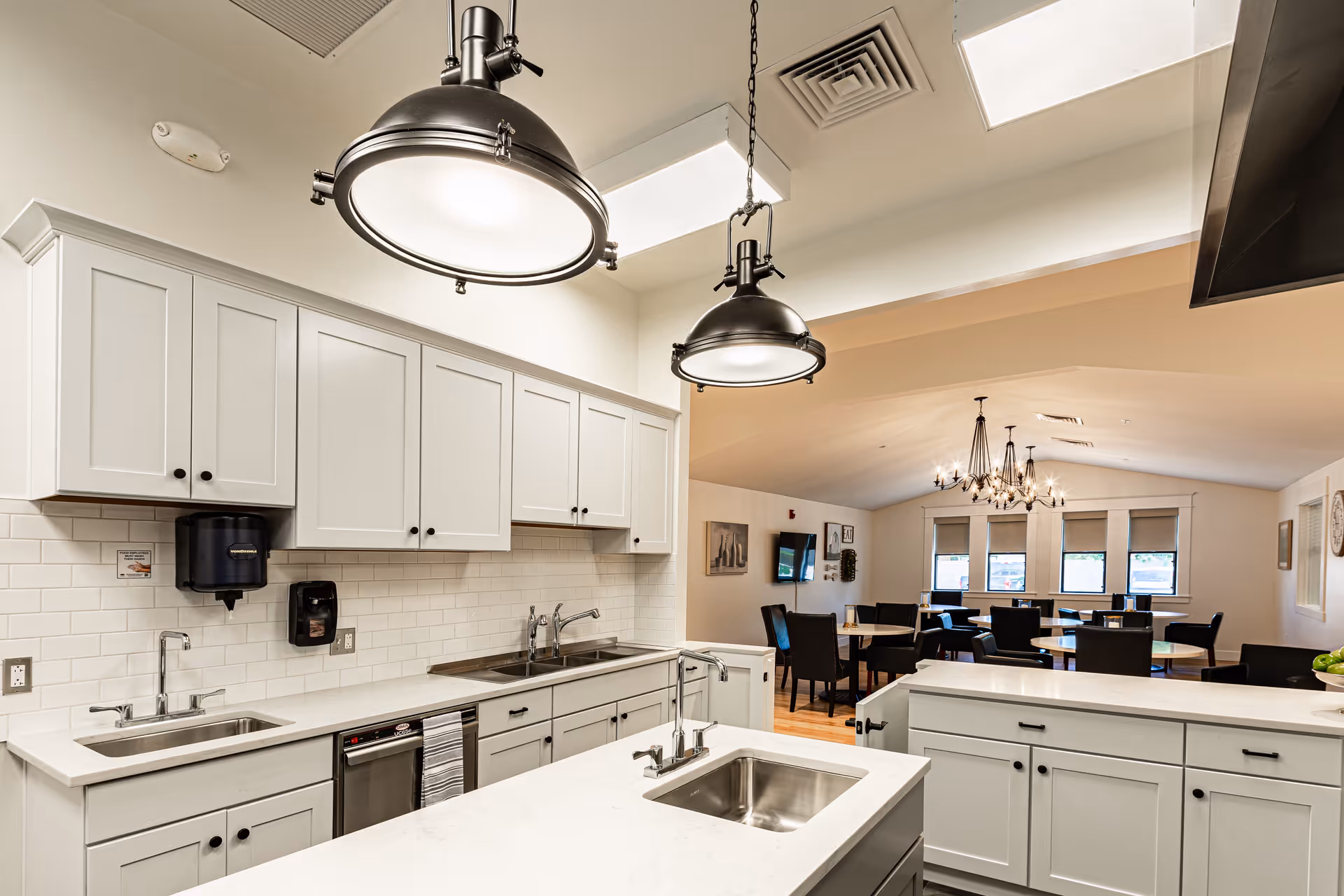 Bright communal kitchen with white cabinetry, two sinks and a central island under industrial pendant lights, opening onto a dining area with tables and chandeliers.