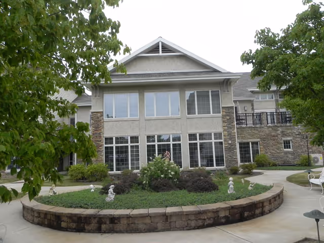Exterior view of a two-story building with large windows and stone accents, surrounded by a landscaped garden with bushes, small statues, and trees. A curved stone retaining wall borders the garden, and a paved walkway encircles it.