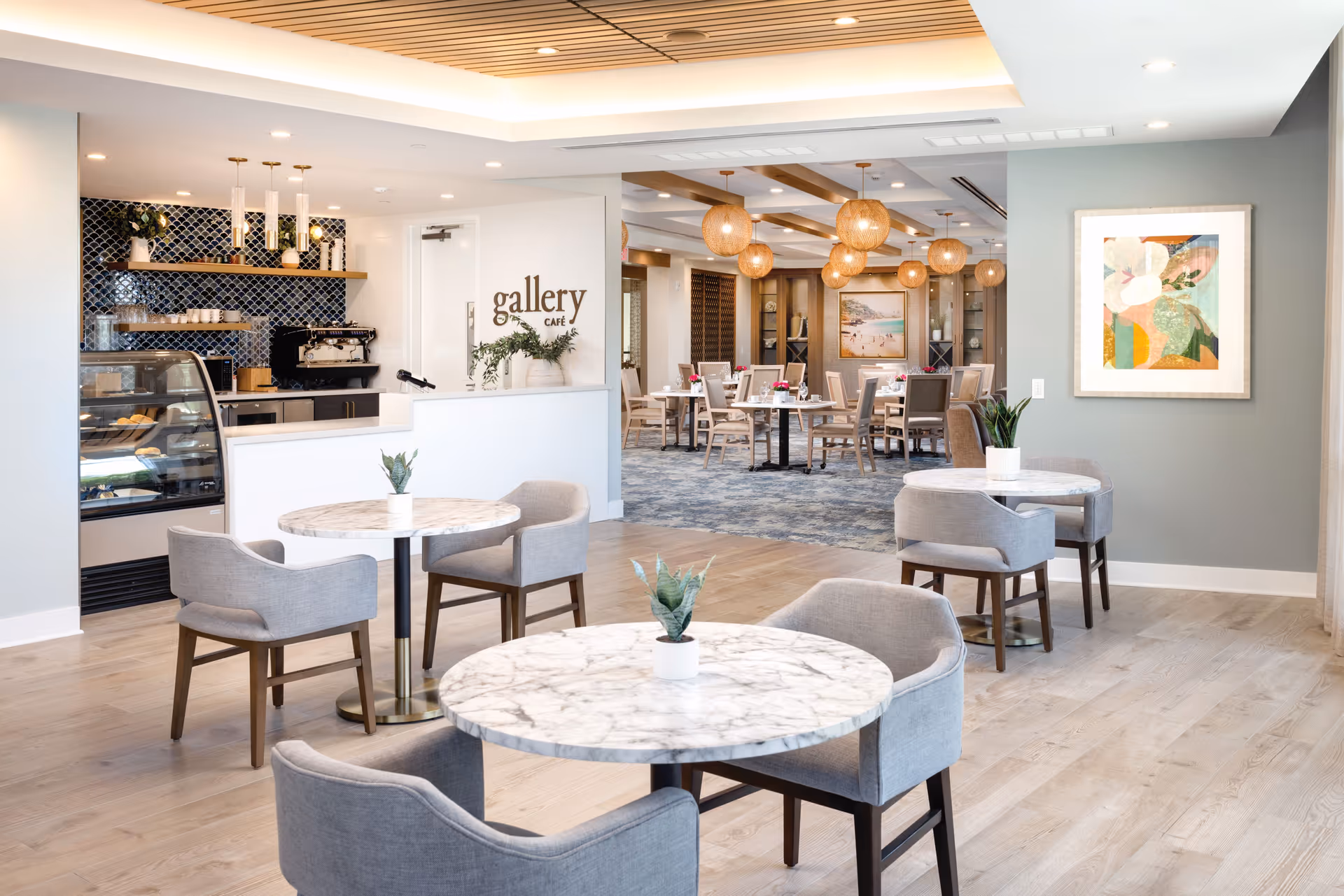 A bright and modern café area with marble-top round tables and gray upholstered chairs. In the background, there is a counter with a display case, coffee machine, and shelves with cups and decor. The wall behind the counter has a dark blue tile backsplash and a sign that reads 'gallery café'. Beyond the counter, there is a dining area with multiple tables and chairs, warm lighting fixtures, and artwork on the walls.