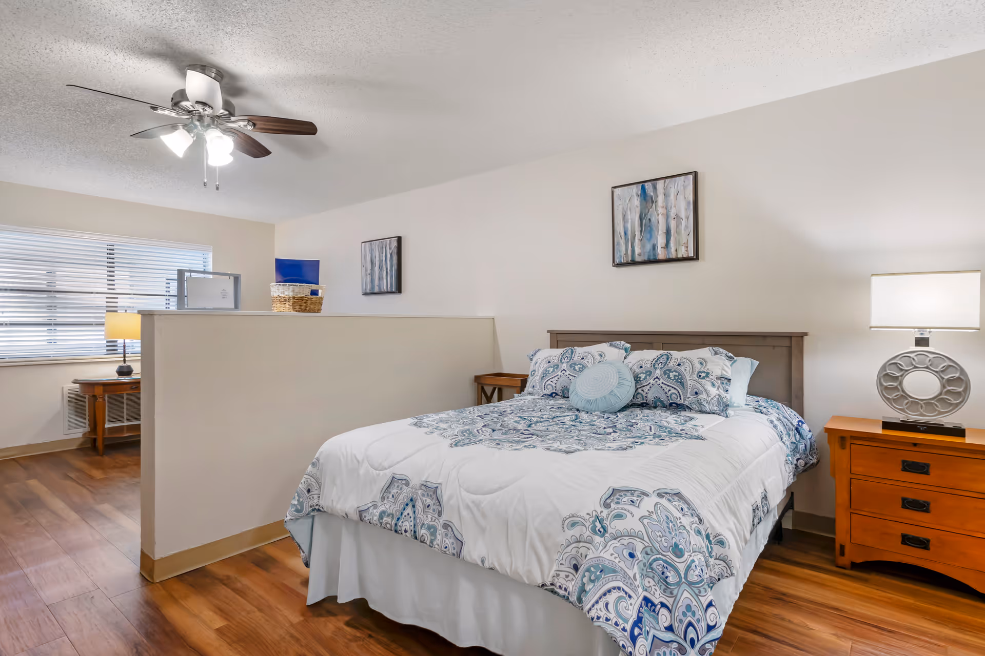 A bedroom with a bed featuring a white and blue patterned comforter and pillows. There is a wooden nightstand with a lamp on the right side of the bed. The room has wooden flooring, a ceiling fan with lights, and two framed pictures on the wall. A half wall separates the bed area from another space with a window and a small table with a lamp.