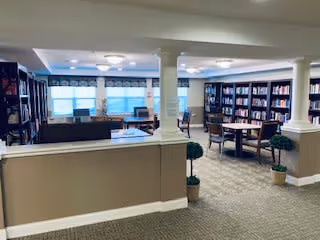 Interior view of a senior living facility library or common area with bookshelves lining the walls, several round tables with chairs, and large windows with blinds allowing natural light to fill the space. The room has beige carpeting and white columns dividing the area.