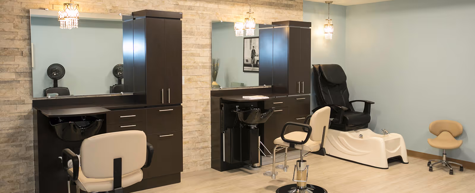Interior view of a salon area in a senior living facility featuring two styling stations with mirrors, black sinks, and beige salon chairs. To the right, there is a black pedicure chair with a white foot bath and a small beige rolling stool. The walls are light blue and beige with a stone accent wall behind the mirrors, and there are hanging light fixtures above each station.