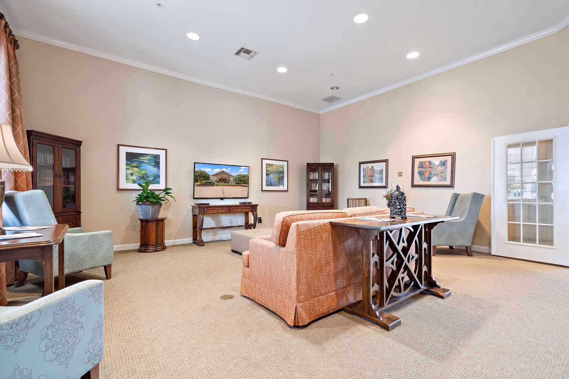 A spacious living room with beige walls and carpeted floor, featuring a peach-colored sofa, two light blue patterned armchairs, a wooden table with decorative items, a flat-screen TV on a wooden stand, framed artwork on the walls, and glass-front cabinets with books and decor.