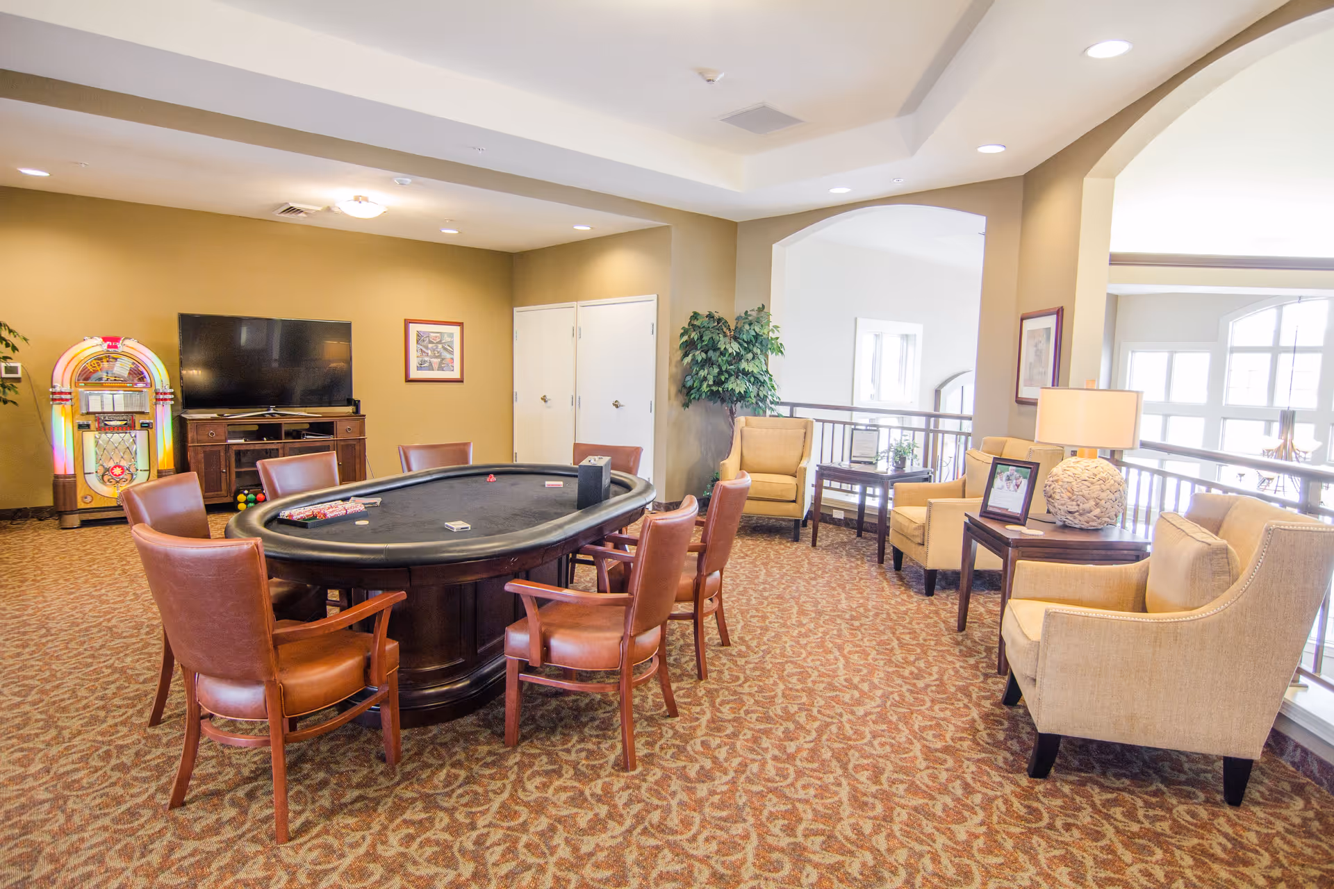 A well-lit common area with a poker table surrounded by brown leather chairs in the center. To the left, there is a jukebox and a flat-screen TV on a wooden stand against a green wall. On the right side, there are beige armchairs with side tables, one holding a lamp and a framed photo. The room has patterned carpet flooring and large windows letting in natural light.
