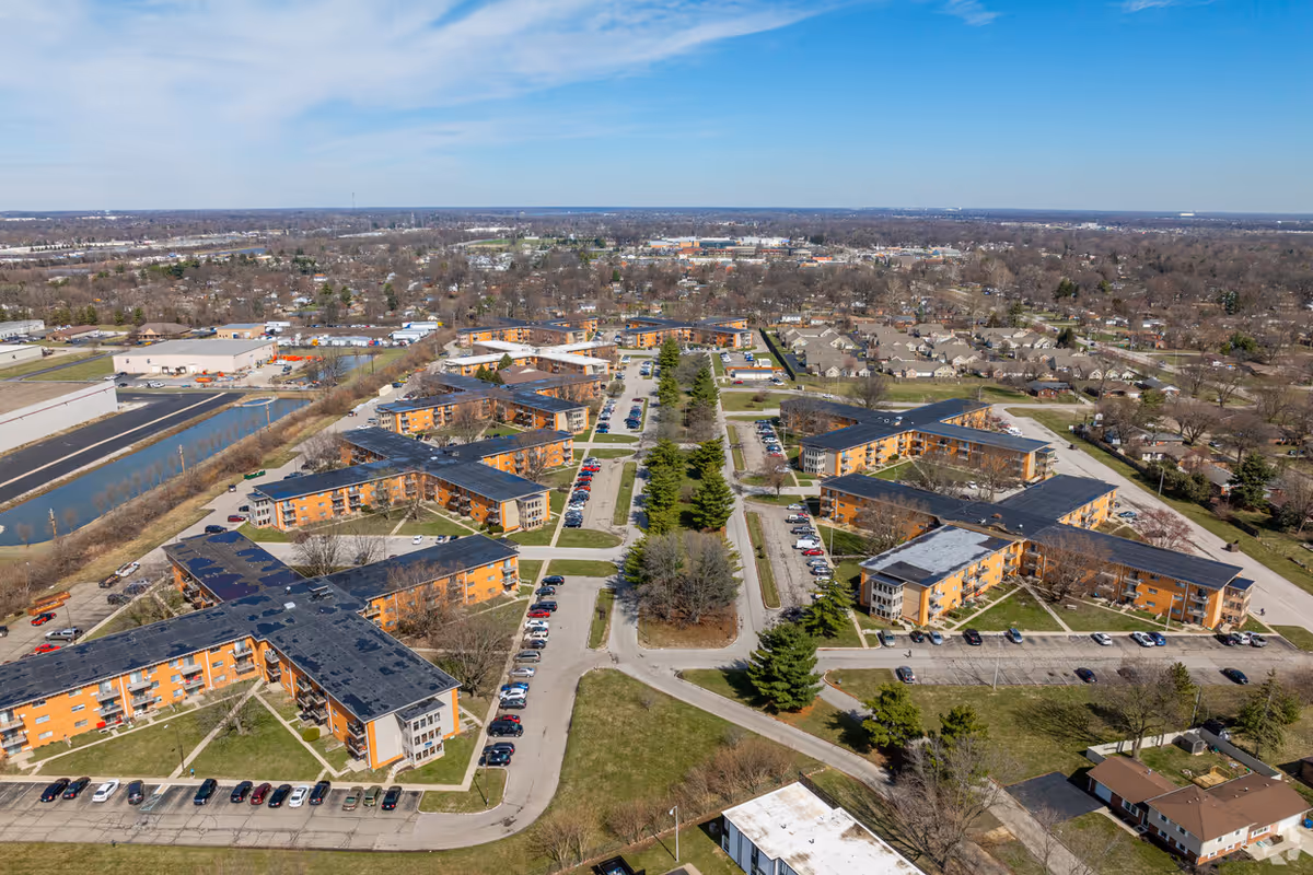 Aerial view of Pinnacle West Apartments, showing multiple orange and beige residential buildings with black roofs arranged around parking lots and green spaces, with a clear blue sky above and a suburban area in the background.