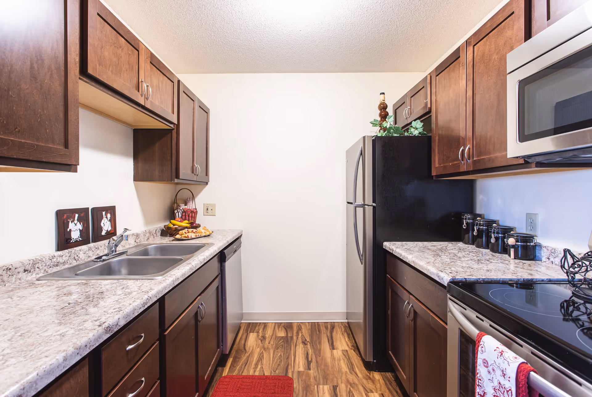 Galley kitchen with dark wood cabinets, marble-look countertops, double sink, stainless steel appliances, and hardwood-style flooring.