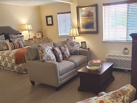 A cozy living and bedroom area in a senior living facility featuring a bed with patterned bedding, a gray sofa with multiple decorative pillows, a wooden coffee table with a bowl and books, two table lamps, framed artwork on the walls, and a window with blinds letting in natural light.
