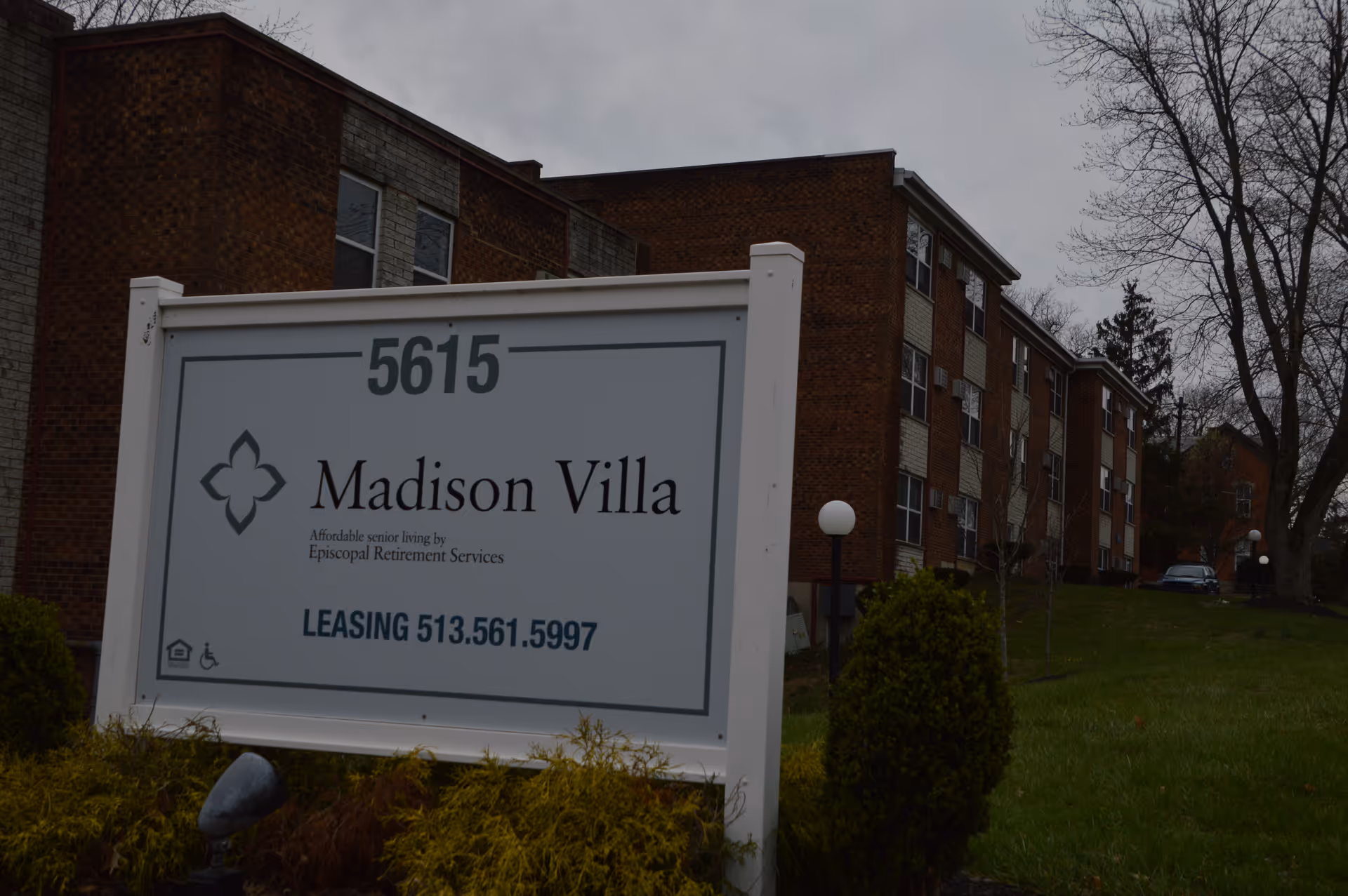 A white sign reading "Madison Villa" and a leasing phone number stands on the lawn in front of a three-story brick senior living building.