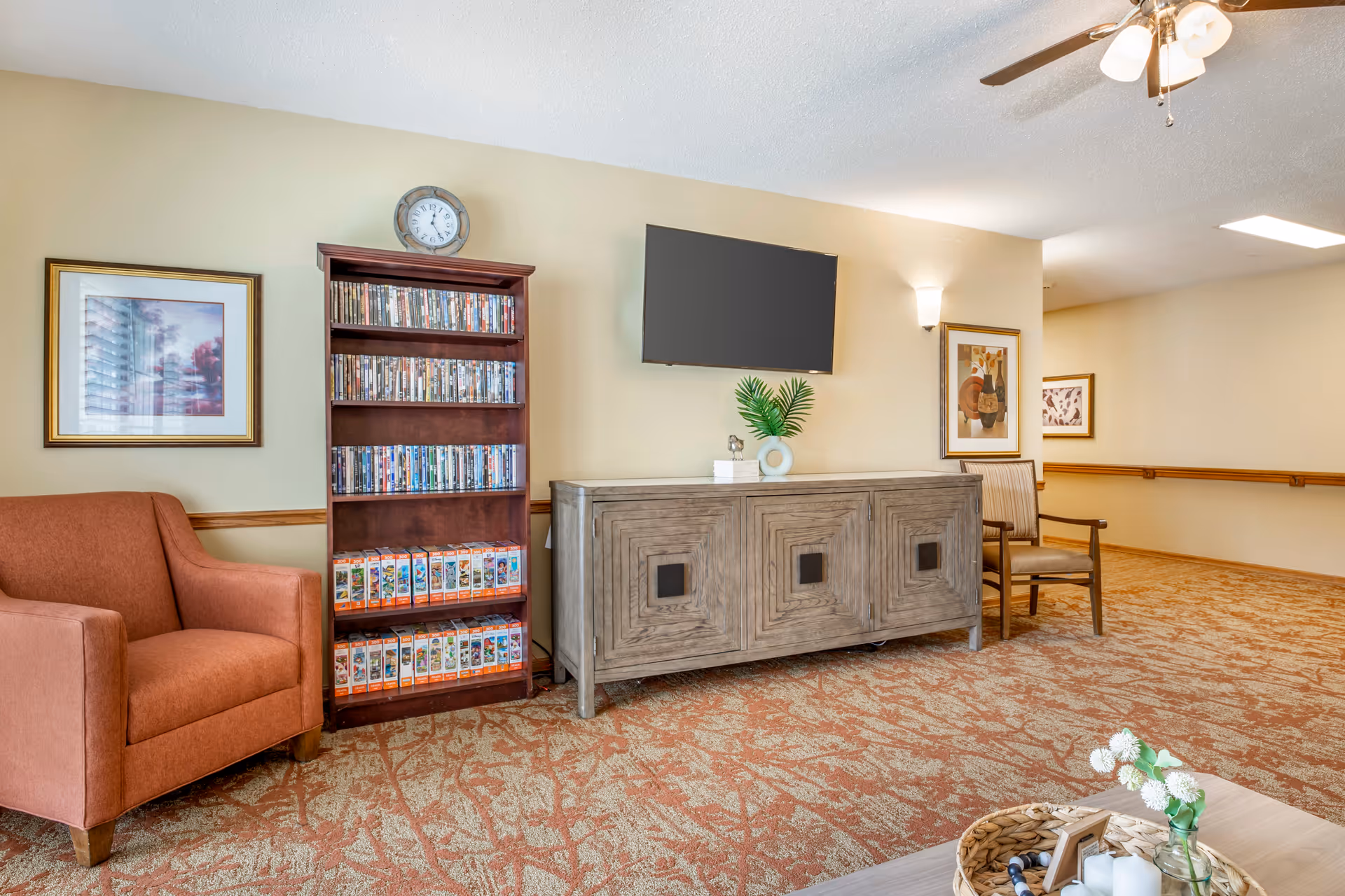 Comfortable sitting area with an armchair, a bookshelf of DVDs, a TV mounted above a wooden cabinet, and decorative accents.