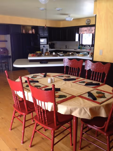 A dining table with a beige tablecloth, rooster placemats and six red wooden chairs in an open kitchen with dark cabinets and yellow walls.