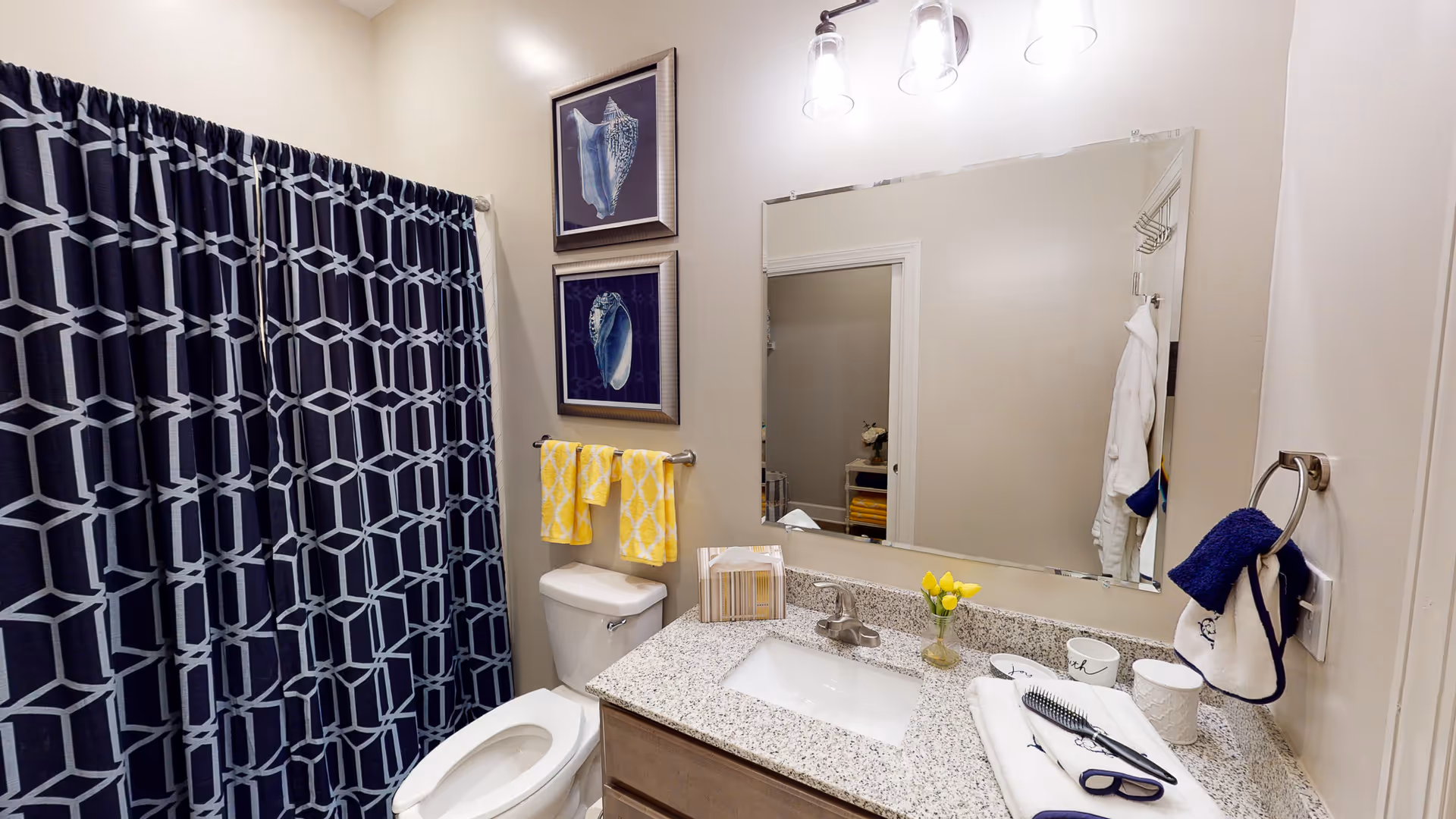 A bathroom with a white toilet, a granite countertop with a sink, and a large mirror above the sink. The shower has a dark blue and white geometric patterned curtain. Two framed pictures of seashells hang on the wall above a towel rack with two yellow towels. On the countertop, there is a tissue box, a small vase with yellow flowers, a hairbrush, folded towels, and some cups. A hand towel hangs on a ring near the light switch.