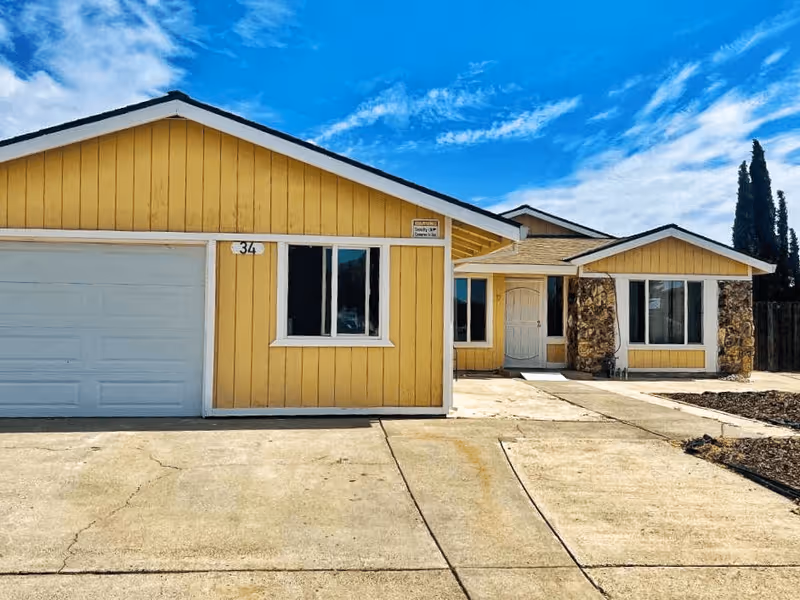Front exterior view of a single-story yellow house with white trim, a garage door on the left, two windows, a white front door, and a concrete driveway under a blue sky with some clouds.