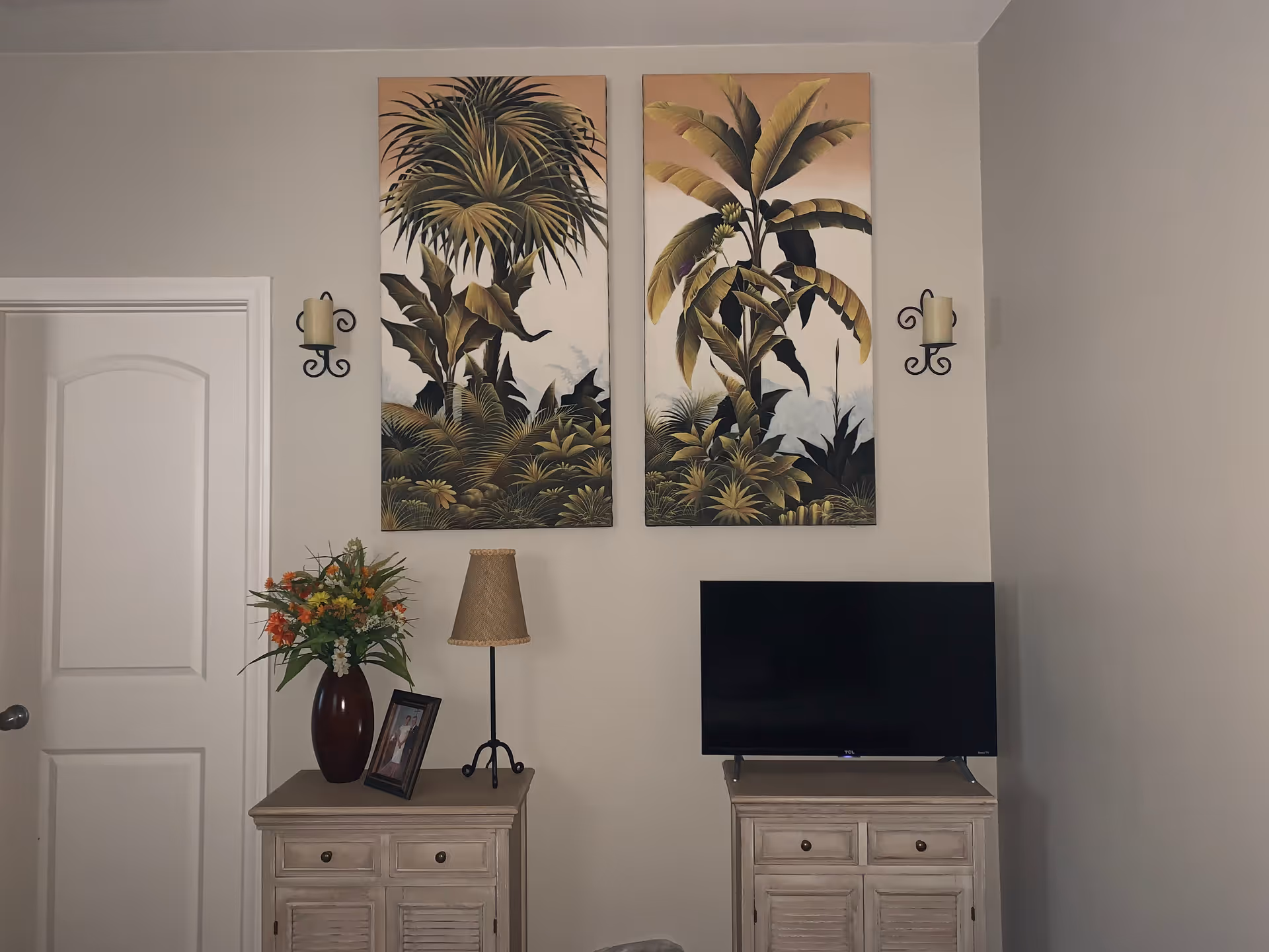 A cozy interior corner featuring two beige wooden cabinets. On the left cabinet, there is a vase with colorful flowers, a framed photo, and a lamp with a woven shade. On the right cabinet, there is a flat-screen TV. Above the cabinets, two large botanical paintings with tropical plant designs hang on the wall. Two decorative wall sconces with candles flank the paintings. A white door is visible on the left side of the image.