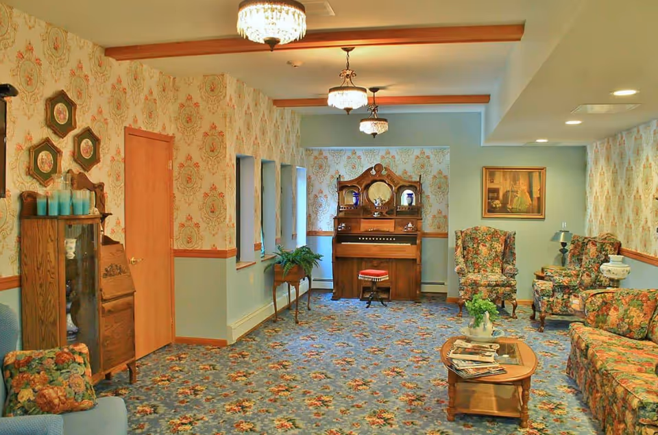 A vintage-style living room with floral patterned carpet and wallpaper. The room features an antique wooden organ with a red cushioned stool, floral upholstered armchairs and sofa, a wooden coffee table with magazines, and decorative wall art. There are three ceiling chandeliers providing warm lighting.