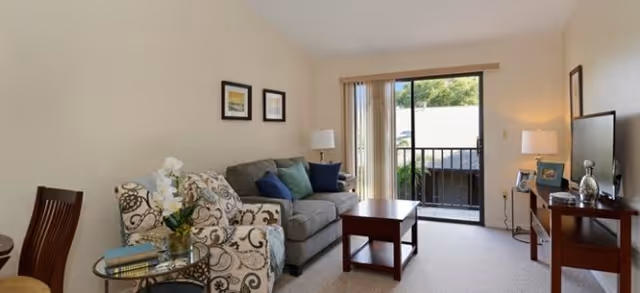 A cozy living room with a patterned armchair and a gray sofa adorned with blue and green cushions. A wooden coffee table sits in front of the sofa. There are two table lamps on either side of the room, one on a side table next to the armchair and another on a wooden TV stand holding a flat-screen television. The room has beige walls and carpeted floor, with sliding glass doors leading to an outdoor balcony with greenery visible outside. Two framed pictures hang on the wall above the sofa.