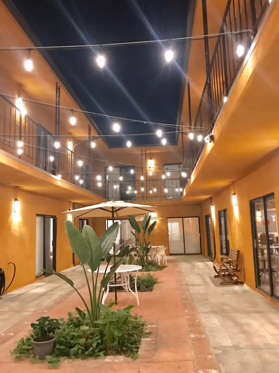 A nighttime view of an outdoor courtyard area in a senior living facility with string lights hanging overhead. The courtyard is surrounded by two-story orange walls with balconies and sliding glass doors. There are plants and small garden beds in the center, white tables with umbrellas, and a wooden bench on the right side.