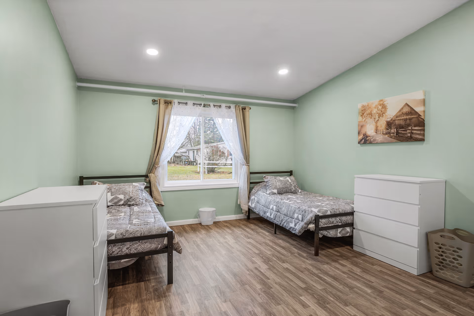 A simple bedroom with two single beds, each with patterned bedding, positioned against opposite walls. Between the beds is a window with sheer white curtains and beige drapes, showing an outdoor view. The room has light green walls, wood flooring, two white dressers, a laundry basket, and a framed picture of a barn on the wall.