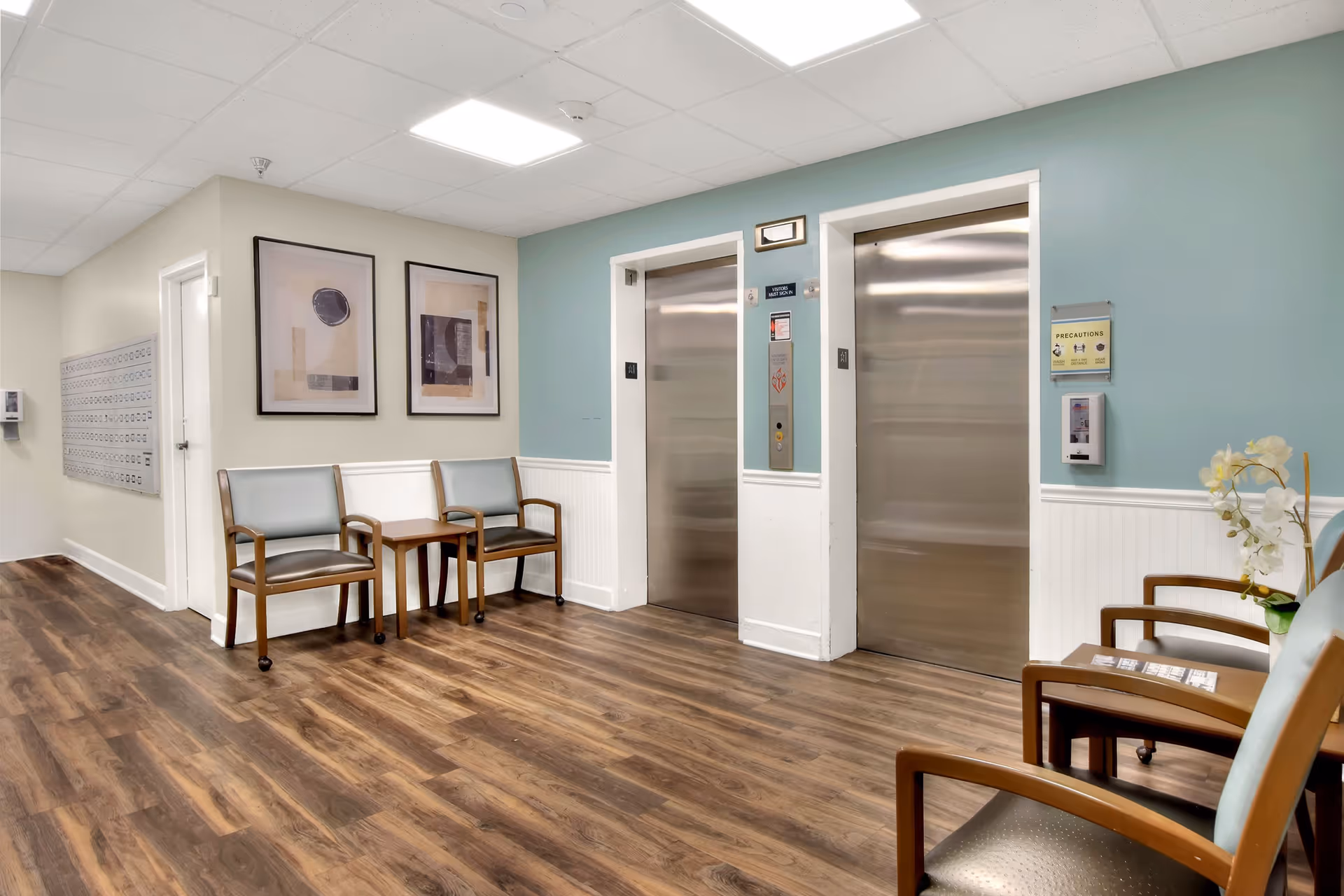 Interior view of a senior living facility hallway with two closed stainless steel elevators, light blue and beige walls, wooden flooring, and several chairs arranged along the walls. There are two abstract framed artworks on the wall and a small table between two chairs. A hand sanitizer dispenser and a sign with precautions are mounted on the wall near the elevators.