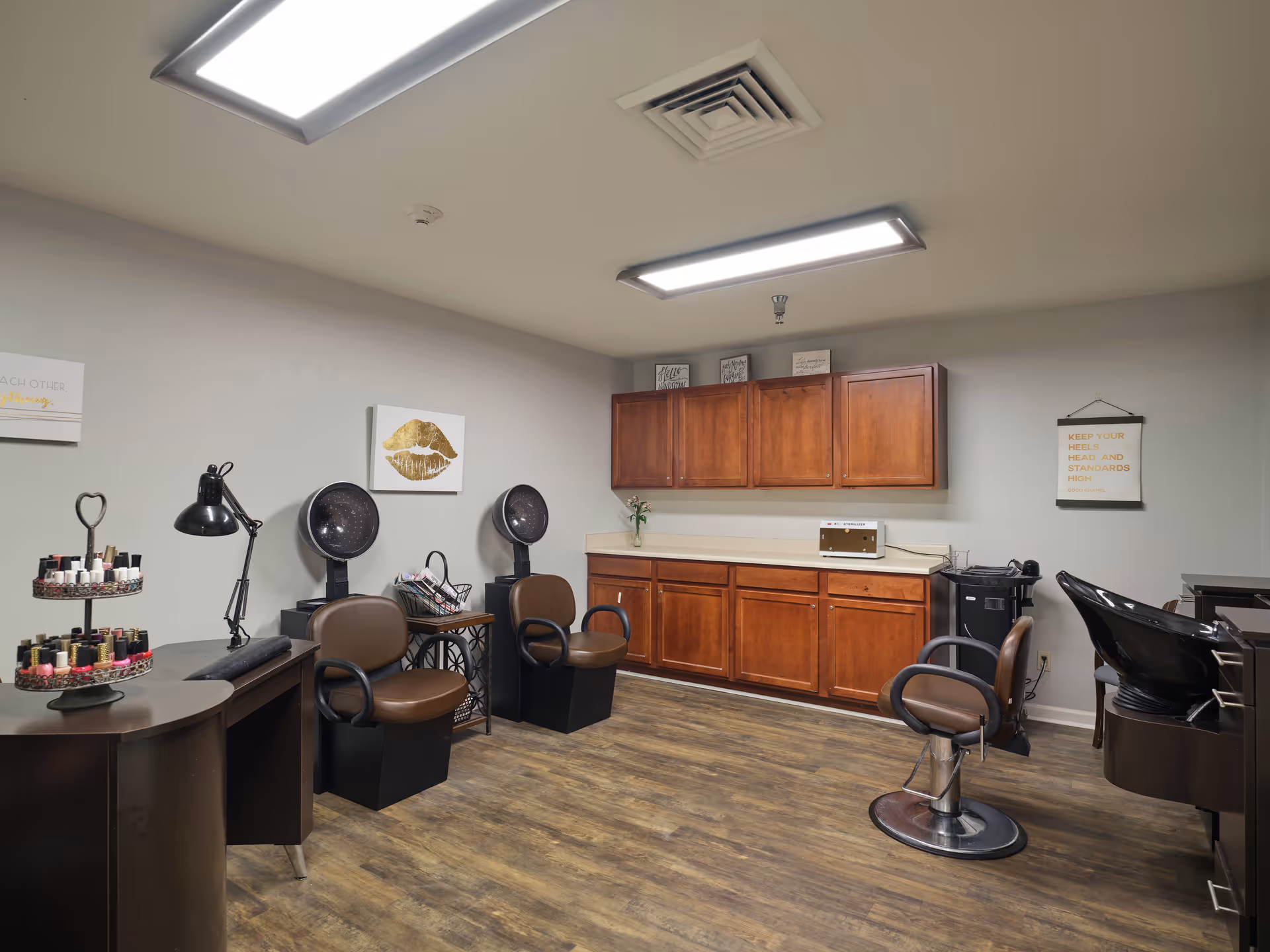 Interior of a salon room with wooden flooring, two brown salon chairs with hair dryers, a styling chair, a black hair washing station, a counter with wooden cabinets above and below, and a display stand with nail polish bottles.