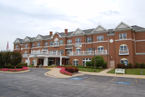 Exterior view of a large three-story brick building with white trim and multiple windows, featuring a covered entrance with columns and a circular driveway. There are landscaped flower beds and bushes near the entrance, a white bench on the lawn, and an American flag on a flagpole to the left.