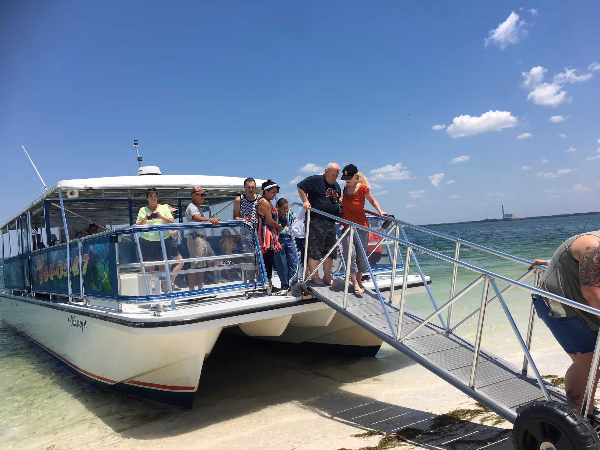 A group of people disembarking from a white catamaran boat onto a sandy beach via a metal gangway under a clear blue sky with scattered clouds.