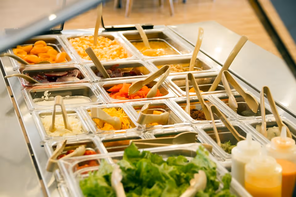 A salad bar with various fresh ingredients including lettuce, shredded cheese, diced onions, sliced carrots, beets, and other vegetables in clear containers with serving utensils. There are also condiment bottles on the side.