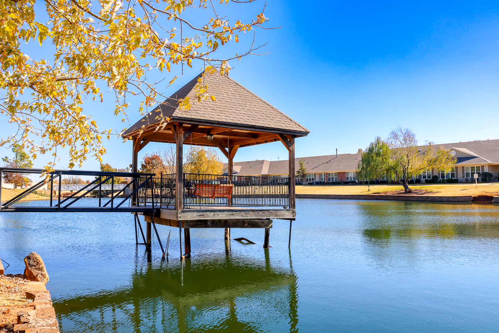 A wooden gazebo with a shingled roof and a swing inside, situated on stilts over a calm pond. The gazebo is connected to the shore by a metal walkway. In the background, there is a large building with multiple windows and a well-maintained lawn with trees under a clear blue sky.