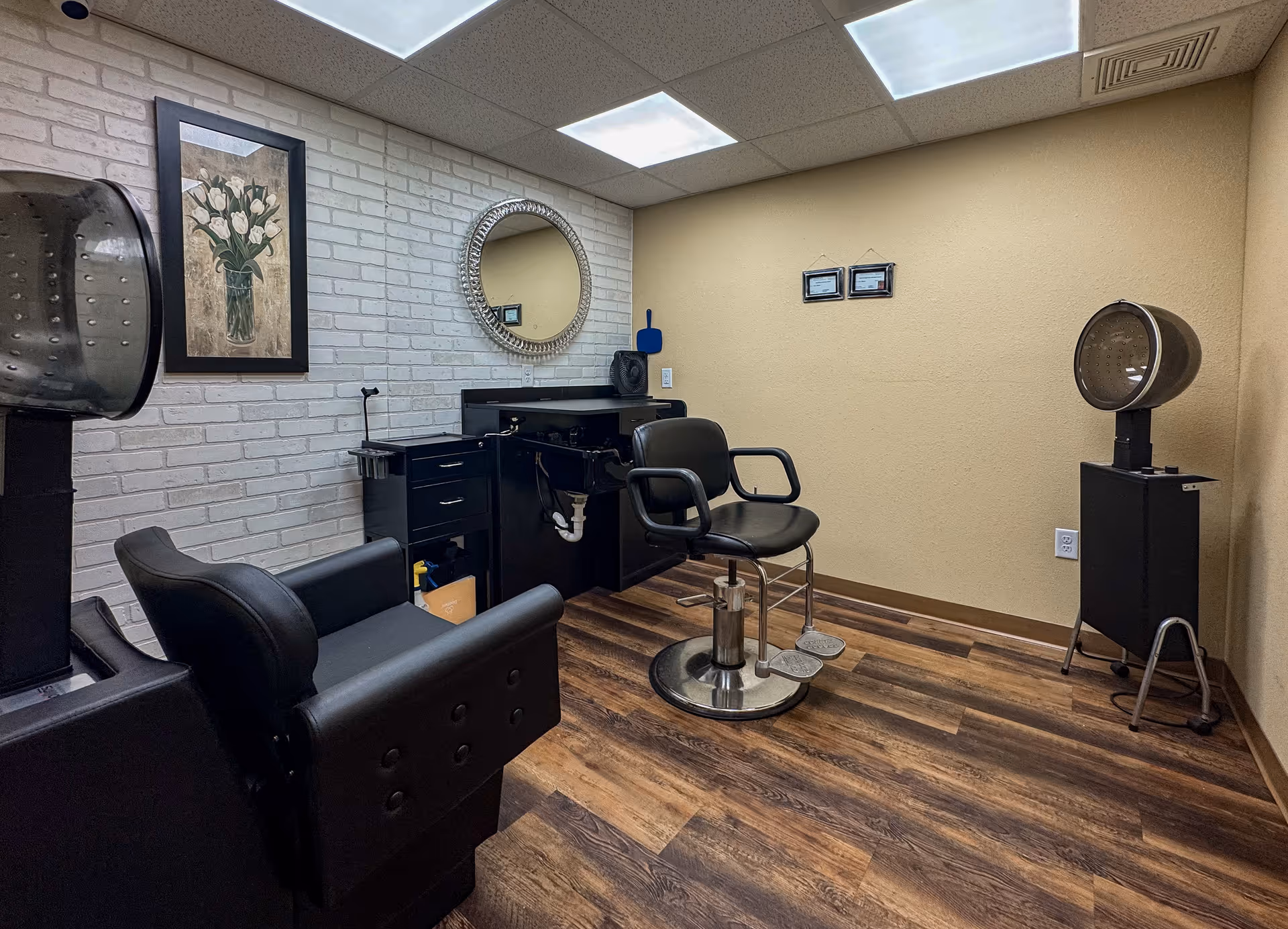 Interior of a small hair salon room with two black salon chairs, a wall-mounted round mirror, a framed picture of white flowers on a brick wall, and two vintage hair dryers on stands. The room has wood flooring and beige walls with ceiling lights.