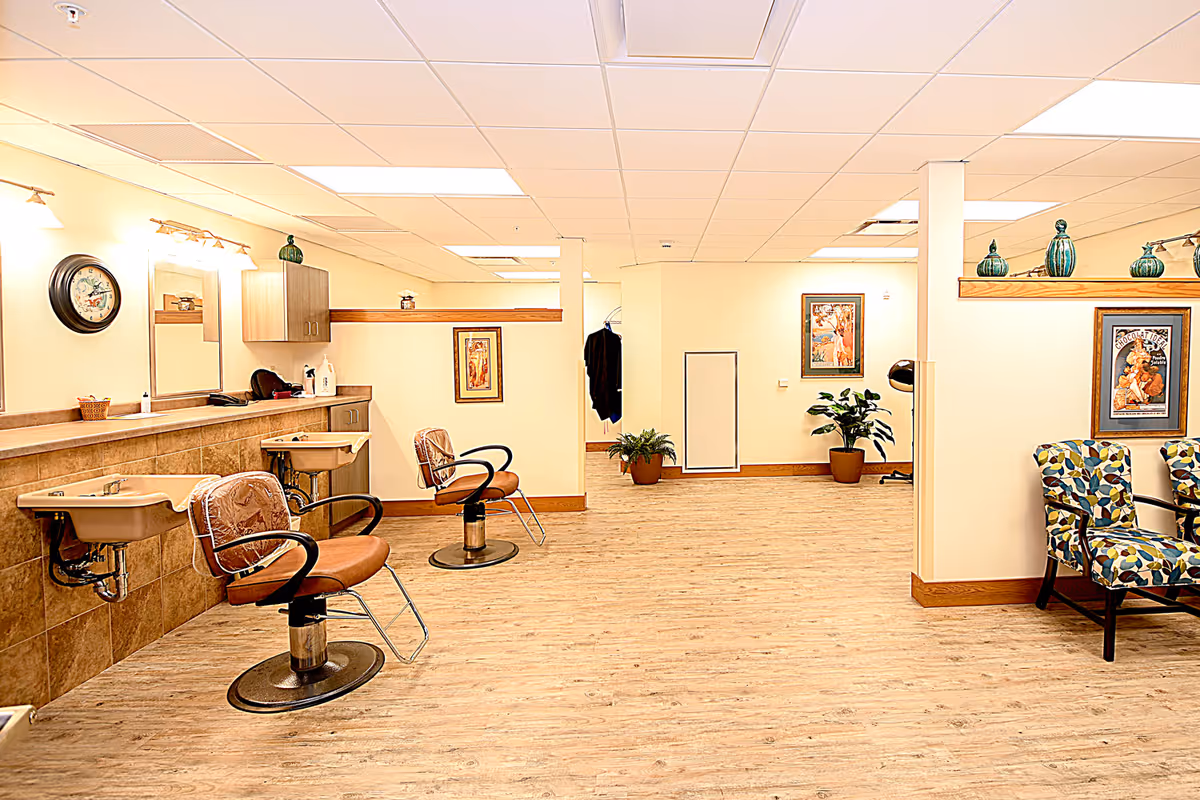 Interior view of a hair salon area in a senior living facility with two salon chairs in front of sinks and mirrors on the left side. The room has light-colored walls, wood flooring, decorative plants, framed artwork on the walls, and patterned armchairs on the right side.